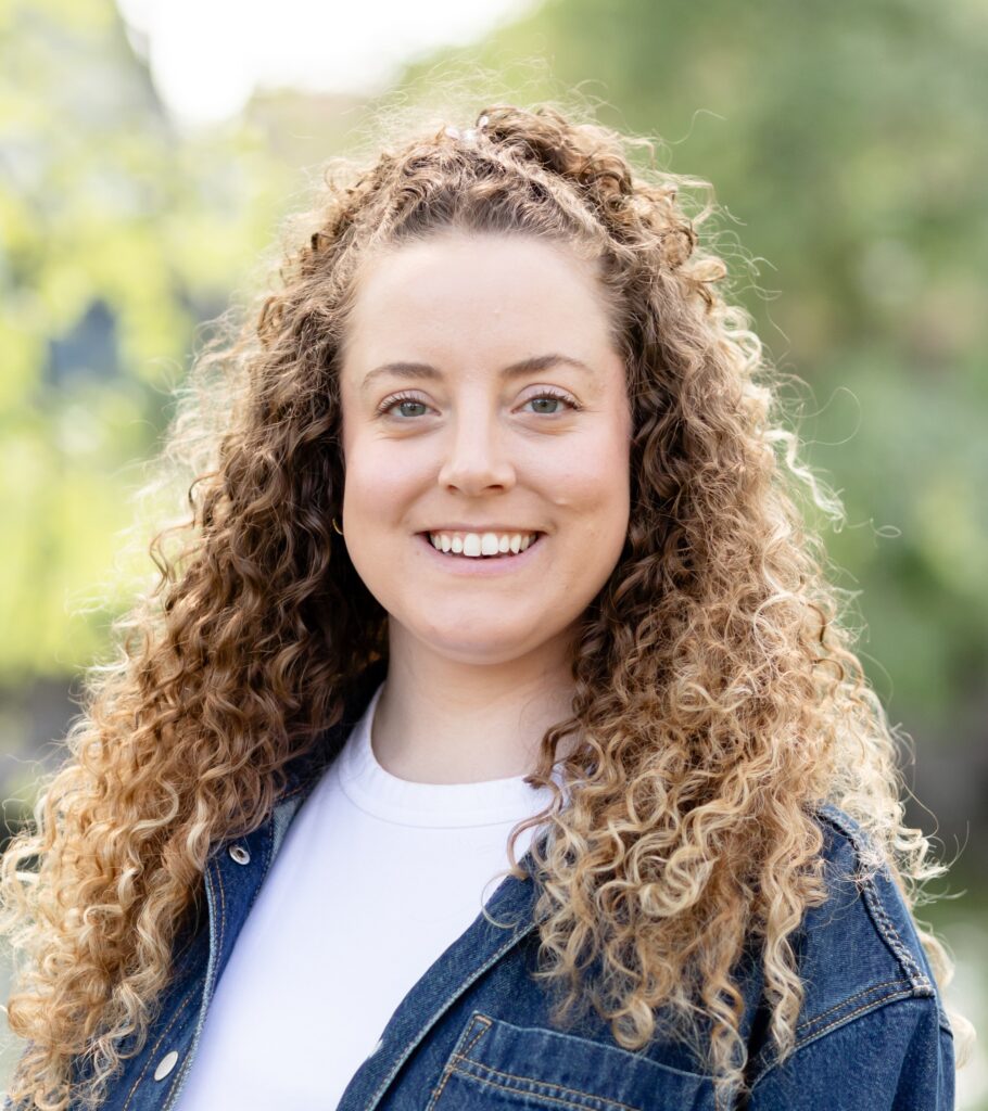 Smiling woman with curly light brown hair wearing a denim jacket outdoors.