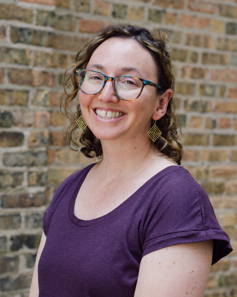 Close-up portrait of a smiling woman wearing colorful tortoiseshell glasses and gold geometric earrings, in a purple top, with a brick wall background.